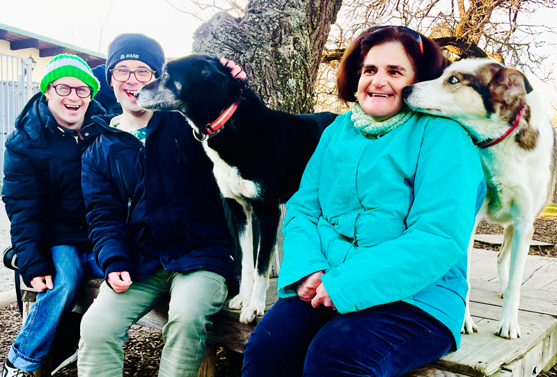 Huskyfieber Menschen mit besonderen Bedürfnisse Eine Frau und zwei Männer mit besonderen Bedürfnissen freut sich über den Besuch bei den Huskys. Sie sitzen auf einer Bank und werden von den Huskys liebkost.